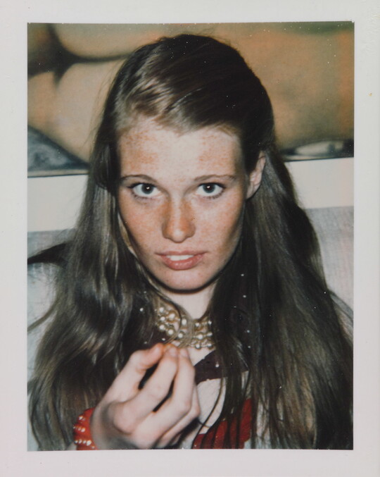 A close-up color portrait photograph of a young White girl with long hair holding something toward the camera.