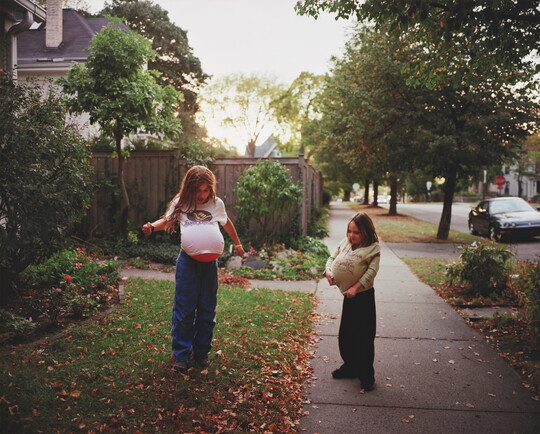A color photograph of two young White girls playing in a front yard with balloons under their shirts mimicking pregnancy.