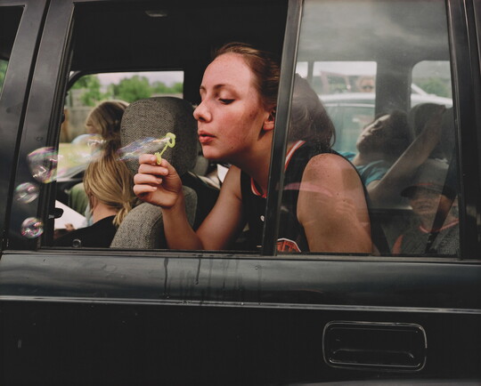 A color photograph of a White teenage girl blowing bubbles out of a backseat car window.