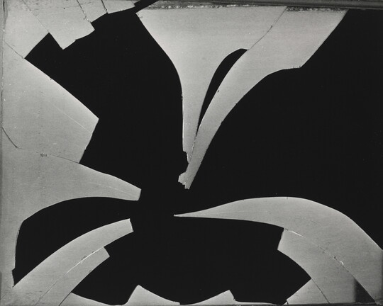 A black-and-white photograph of a pane of broken glass with many pieces missing in front of a black background.