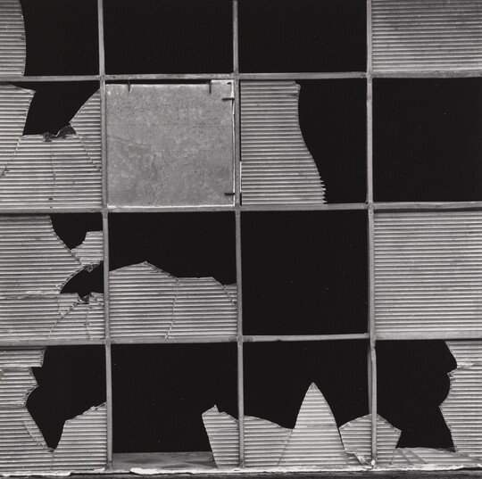 A black-and-white photograph of a metal grid with broken or missing textured glass pieces in each section.