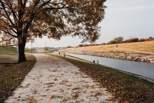 A color photograph of a late fall landscape with a river disappearing into the distance and people running on well-groomed paths.
