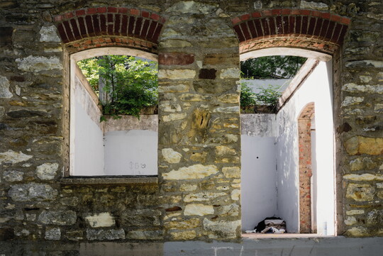 A color photograph of two brick arches, one a window the other a door, in a stone wall, looking into an empty, white room with no roof and green vegetation overhead.