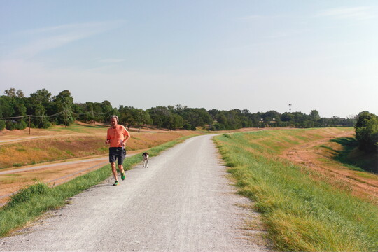 A color photograph of a middle-aged White man and an unleashed dog running toward the viewer on a paved path.