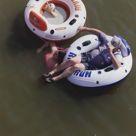 A color photograph taken from above of a White man reclining in an innertube and a White woman in pink swimsuit soaking in the water next to an innertube.