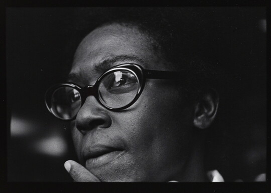 A black-and-white close-up photograph of the face of a Black woman wearing black glasses, an index finger held next to her mouth.