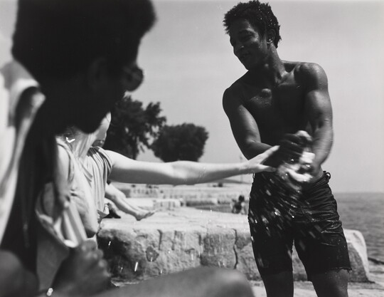A black-and-white photograph of a smiling Black man in swim shorts wringing water out of something as a White woman reaches toward him.