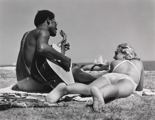 A black-and-white photograph of a Black man playing guitar on a beach blanket next to a White woman lying on her stomach with her feet toward the camera.