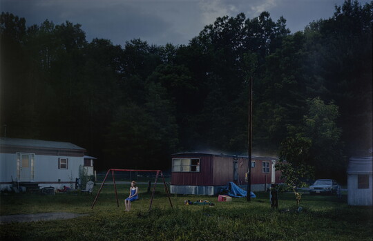 A color photograph of a White teenage girl sitting on a swing set in front of a mobile home park.