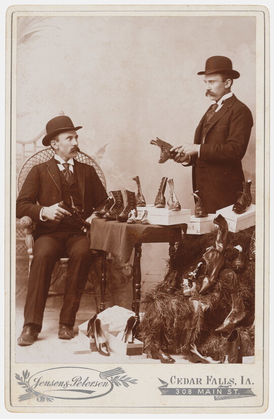 A black-and-white studio photograph of a seated White man in a bowler hat looking at shoes as the same man standing appears to show him shoes.