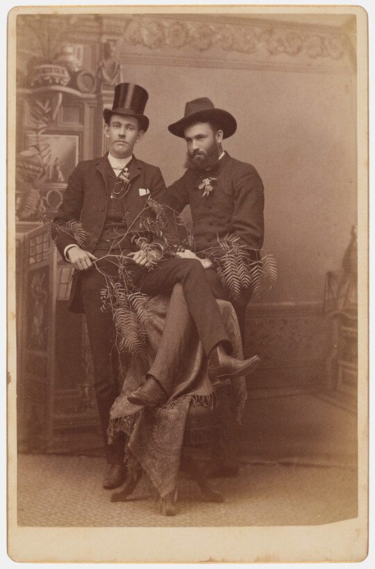 A sepia-toned studio photograph of two White men in suits, each standing with one a leg over the back of a single chair so that their legs overlap.