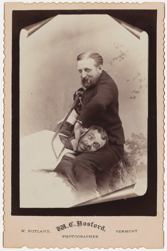 A sepia-toned studio photograph of a seated White man holding a saw against the neck of another White man whose head is resting on his lap.