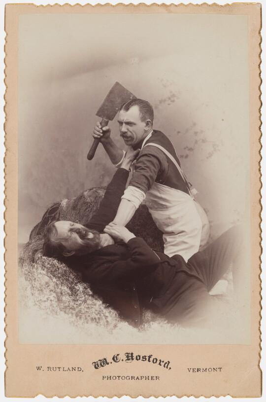 A sepia-toned studio photograph of a White man in a butcher's apron holding a cleaver with one hand as he holds by the neck another White man who is reaching up toward the cleaver.