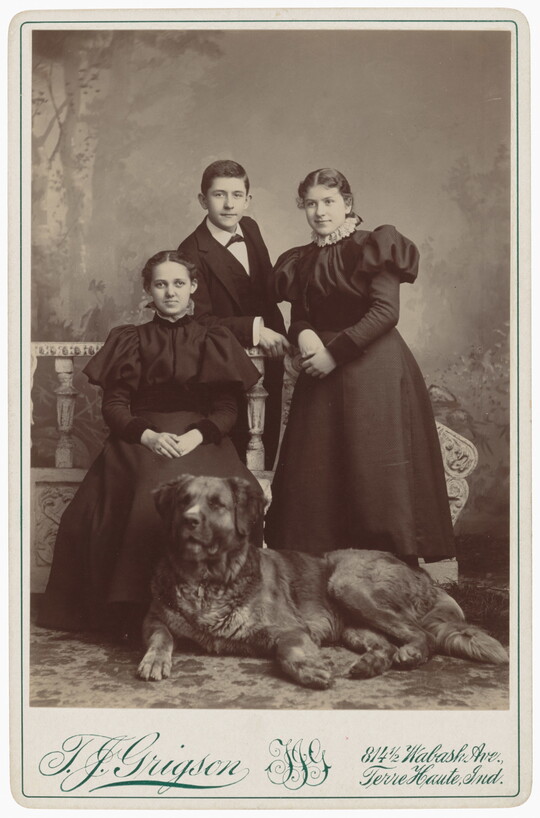 A black-and-white studio portrait photograph of three young adults, two White women and one White man, posing behind a large dog.