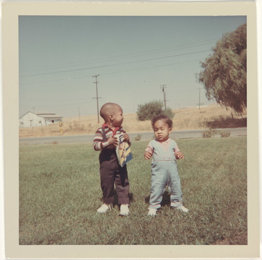 A color photograph of two Black toddlers standing in a grassy yard.