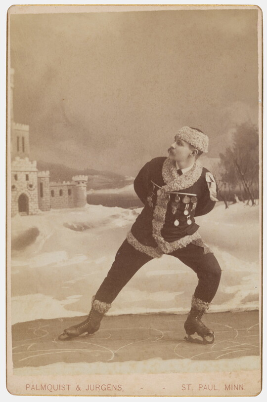 A sepia-toned studio portrait photograph of a White man dressed in winter clothes pretending to ice skate.