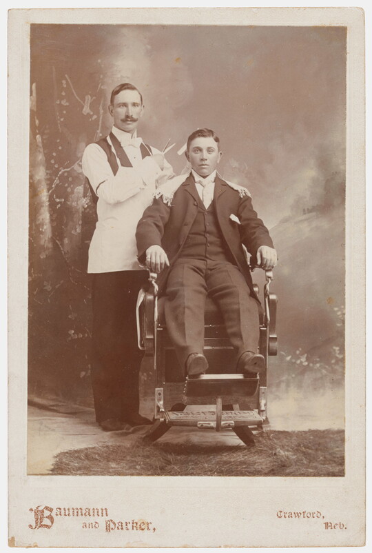 A sepia-toned photograph of two White men in Victorian clothing, one seated in a barber's chair, the other standing behind the chair holding scissors.