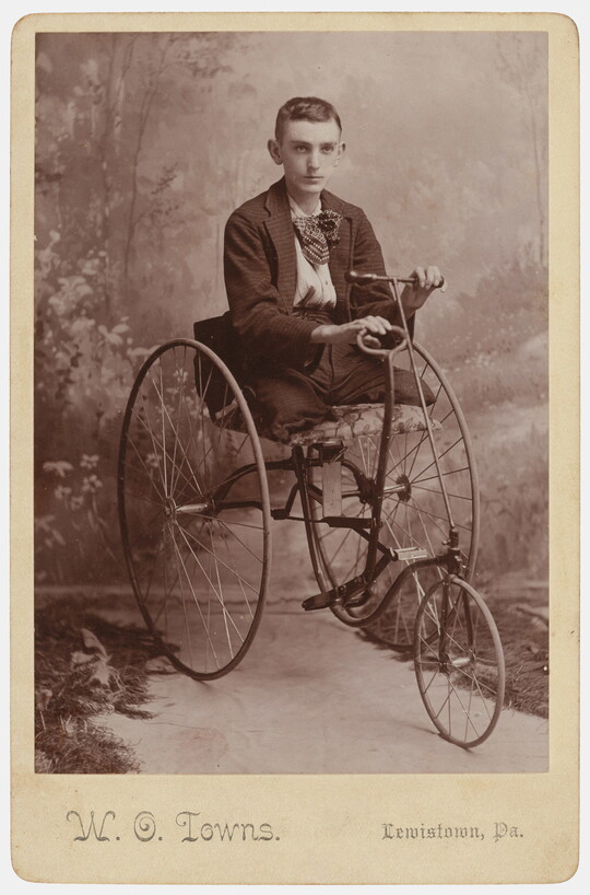A sepia-toned studio photograph of teenage White boy with no legs, seated in a Victorian tricycle-style wheelchair.