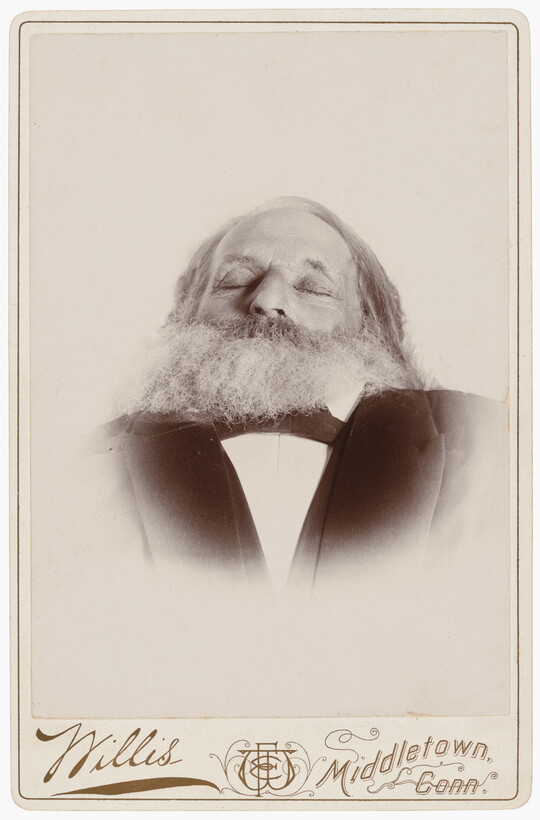 A black-and-white photograph of a deceased White man, from the shoulders up, with a full beard and eyes closed wearing a suit.