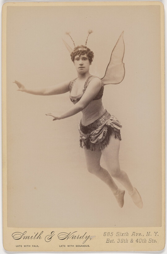 A sepia-toned studio photograph of a White woman dressed as a fairy, including wings and antennae, suspended in the air against a white background.