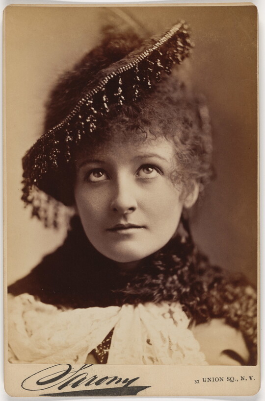 A sepia-toned portrait photograph of a young White woman with curly hair, eyes looking up toward her beaded hat.