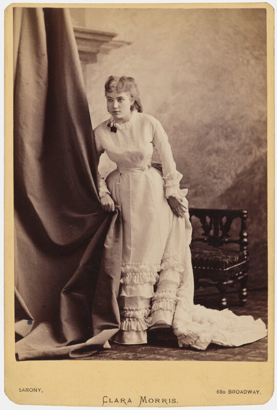 A sepia-toned studio portrait photograph of a young White woman in a long dress with ruffles along the bottom standing next to a chair and clutching a curtain.