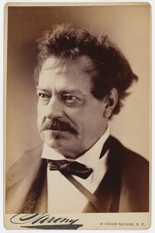 A sepia-toned studio portrait photograph of a middle-aged White man with unkempt hair, a mustache, wearing a black jacket, white shirt with a high collar, and bow tie.