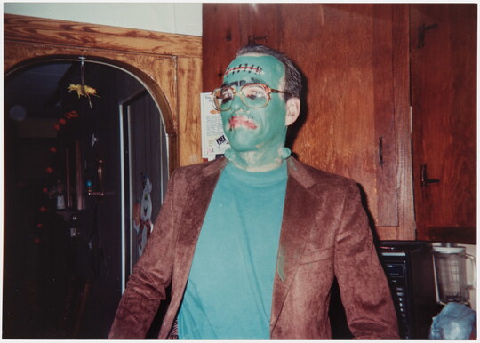 A color photograph of a man wearing green face paint and a Frankenstein costume standing in what looks like a kitchen.