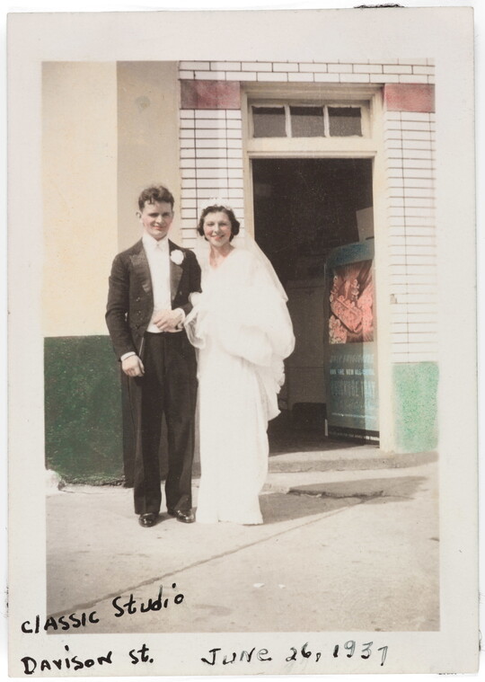 A color photograph of a young White man and woman dressed in wedding attire and standing in front of a doorway.