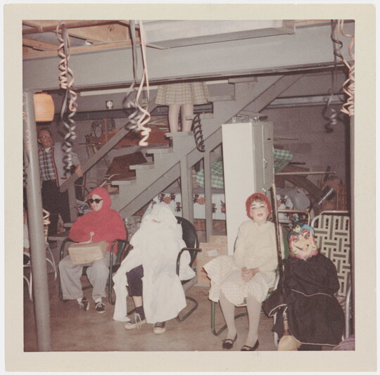 A faded color photograph of four children in costumes sitting in chairs at the foot of basement stairs.