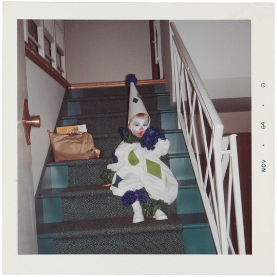 A color snapshot of a toddler in a blue, green, and white clown costume sitting on stairs inside a house.