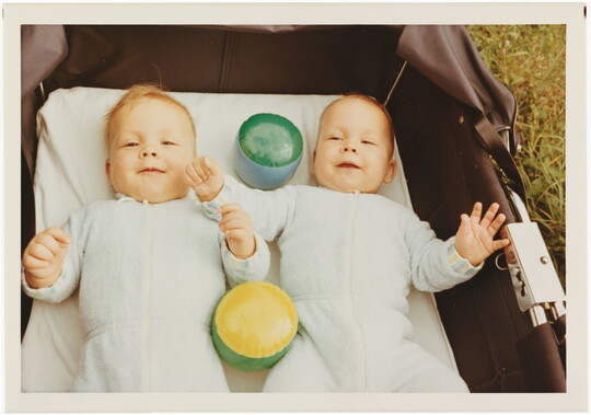 A color photograph of two White babies smiling and lying on their backs with toys in a baby carriage that is sitting in grass.