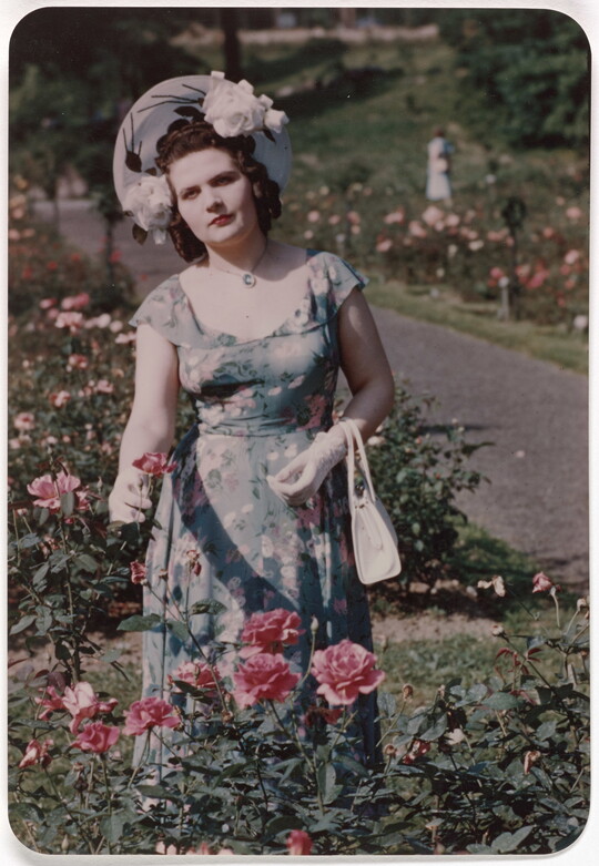 A color photograph of a White woman wearing a sunhat, floral dress, and white gloves standing in a rose garden.