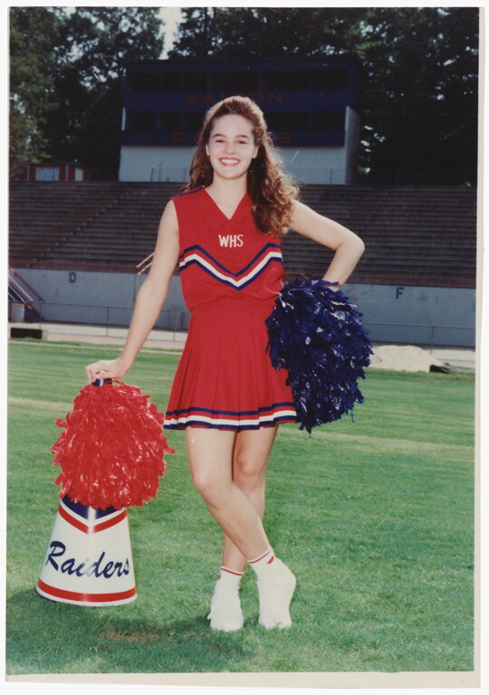 A color photograph of a young, smiling White woman in a red cheerleader uniform, holding pom-poms and leaning on a megaphone on a grassy sports field.