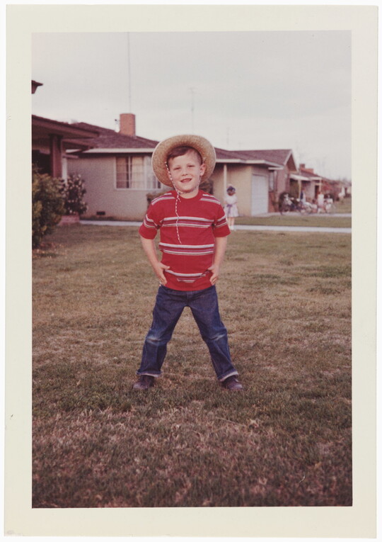 A color snapshot photograph of a young White child standing in a yard wearing a cowboy hat, red striped shirt, and blue jeans.