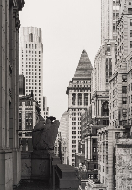 A black-and-white photograph of a cityscape taken from a balcony with a sculpted eagle atop the corner of the balustrade, skyscrapers rising around it.