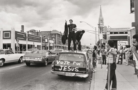 A black-and-white photograph of a downtown street scene of a man preaching from the top of a car with "Vote for Jesus" spray-painted on the back.