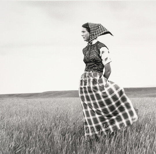 A black-and-white photograph of a young White woman wearing a long plaid skirt and a headscarf standing in field.