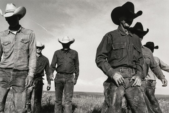 A black-and-white photograph of six cowboys walking in a field toward the viewer with their faces in shadow from their cowboy hats.