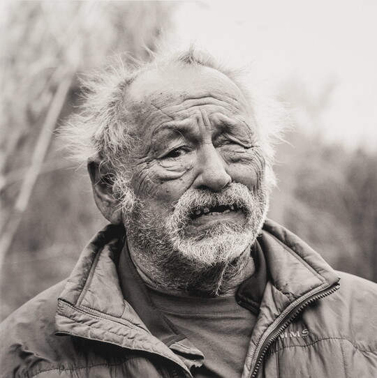 A black-and-white portrait photograph of a older light-skinned man, balding and with overgrown facial hair, wearing a quilted jacket standing outdoors.