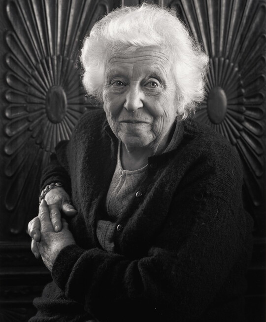 A black-and-white portrait photograph of an elderly White woman with fluffy white hair, posed in front of a carved wooden wall.