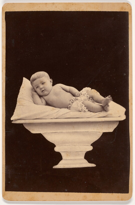A black-and-white studio portrait photograph of a White baby in a diaper reclining on a pillow placed on a pedestal.
