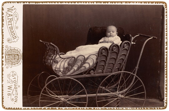 A black-and-white studio portrait photograph of a bald White baby posed on white pillows in an ornate baby buggy.