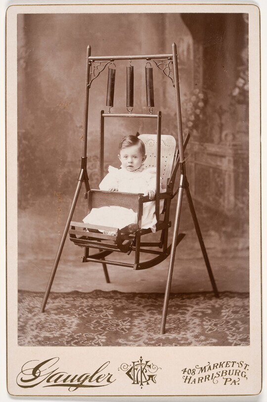 A black-and-white studio portrait photograph of a White toddler with dark hair sitting in a metal swing with three springs supporting the chair from the swing frame.