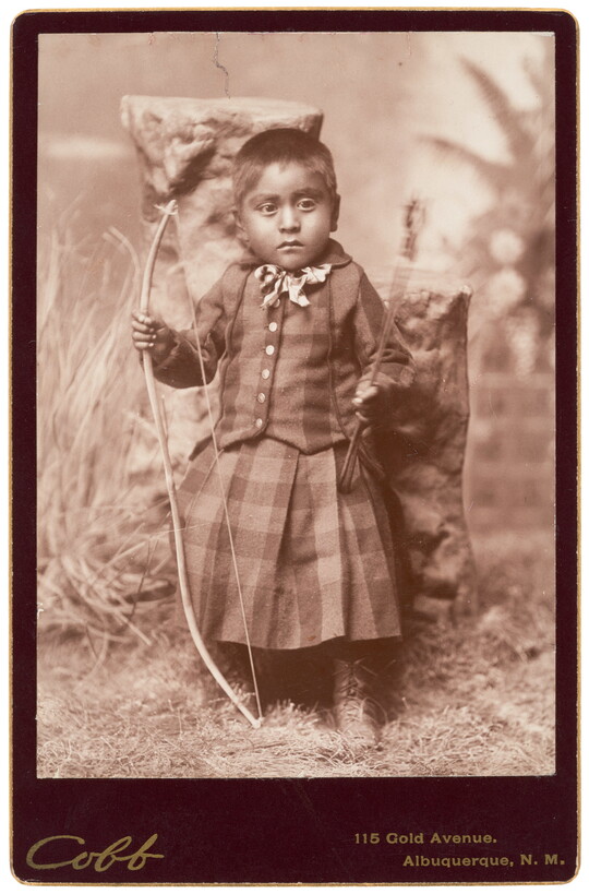 A sepia-toned studio portrait photograph of a young medium-skinned child with closely cropped dark hair wearing a tartan outfit and holding a bow and arrow.