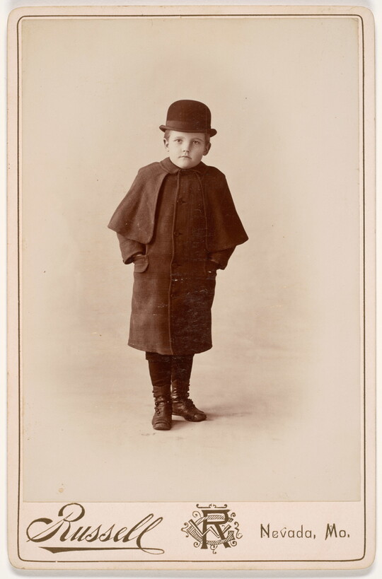 A black-and-white studio portrait photograph of a young White boy wearing a bowler hat, hands tucked into the pockets of a coat with a cape.
