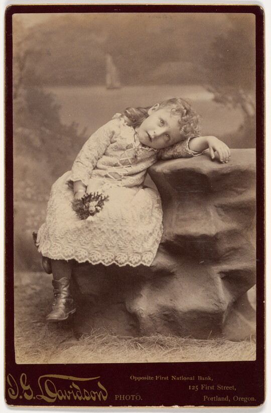 A black-and-white photograph of a young White girl with curly, light-colored hair in a lace dress, draped on a fake rock, with her booted feet dangling.