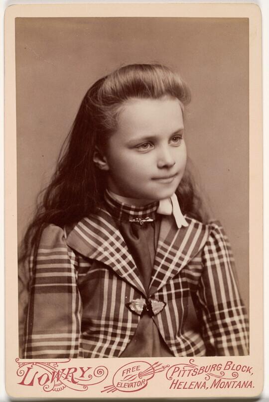 A black-and-white studio photograph of a young White girl with long hair wearing a plaid long-sleeved jacket.