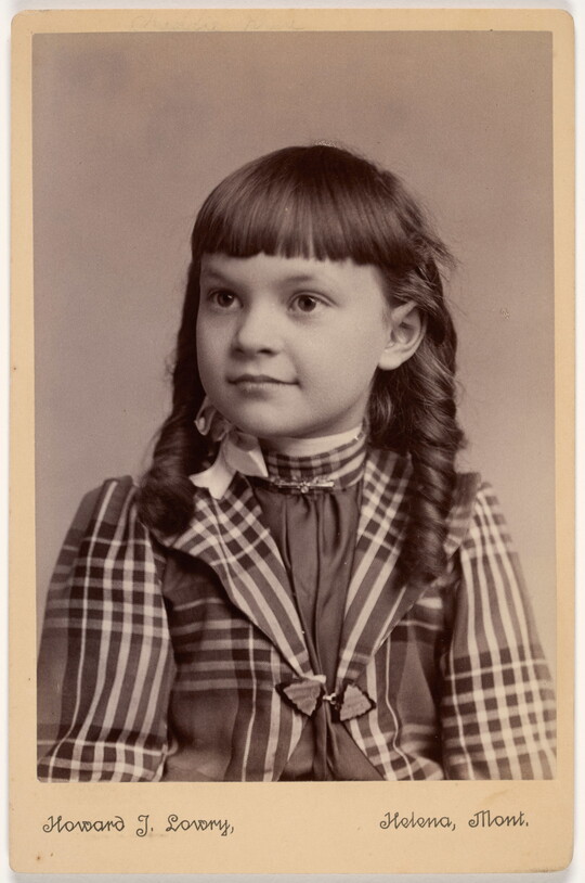 A black-and-white studio photograph of a young White girl with bangs and long hair styled in banana curls wearing a plaid jacket.