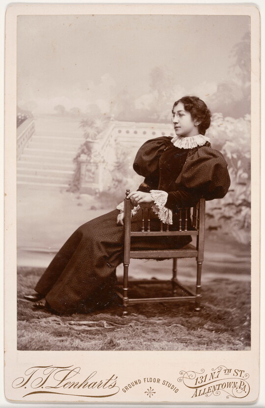 A black-and-white studio photograph of a young White woman seated in profile wearing a high-necked gown with puffy sleeves and lacy cuffs.